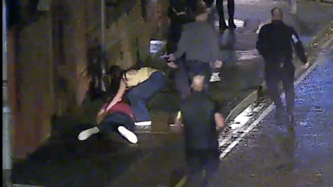 CCTV image of the victim lying face down in the street, a woman is crouched over him. Security staff are looking up the street.