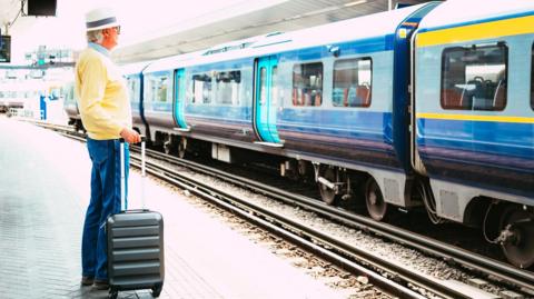 A senior male in his 70s waits at the station platform, ready to go on holiday. He has a small wheelie suitcase and wears a summer hat. He has a pair of headphones around his neck. On the opposite platform, a blue Southeastern train waits to leave.