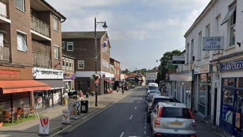 A Google Streetview image showing High Street in Ewell which has cars parked along one side and shops on either side. IN the distance the road bends out of site and more buildings can be seen