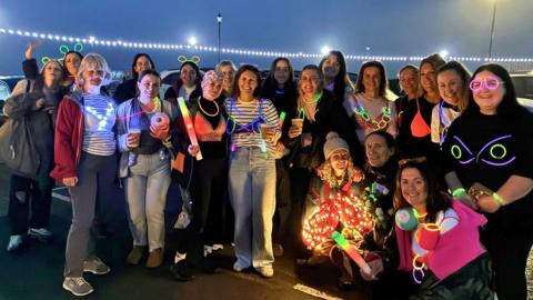 A group of about 20 people gather in a dimly-lit car park in the evening, wearing glow-in-the-dark accessories like necklaces, bracelets, and glasses. The women wear bras over the top of their clothes.