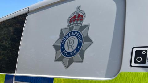 A silver, blue and red Humberside Police logo on the side of a van. The logo features a crown, royal emblem and the words "Humberside Police". The van is white with yellow and blue graphics.