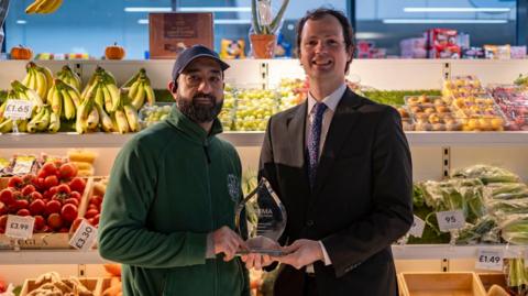 A fruit and veg stall holder holds the award with Alex Ross Shaw. The trader is wearing a green fleece and blue hat, while Ross Shaw is in a black suit.they are stood in front of a market stall selling fruit and vegetables, including bananas, tomatoes and grapes 