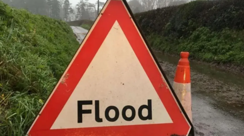 A triangular sign reads 'Flood', a cone stands behind it on a dirt track road with grassy hills at either side 