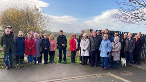 A group of people standing together outdoors on a paved area near a wooden fence and a leafless tree. They are dressed in winter coats of various colors, including red, blue, gray, and beige. The background shows a grassy field and trees with autumn foliage under a partly cloudy sky.