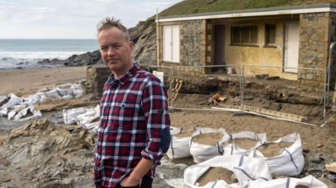 Mullion Surf Life-Saving Club representative Simon Mitchell stands on Polurrian Beach with the former lifeguard hut behind him. There are a lot of bags of building sand dotted in front of the building and metal fencing surrounding the hut. It is a cloudy day and Simon is wearing a red, white and blue checked shirt.