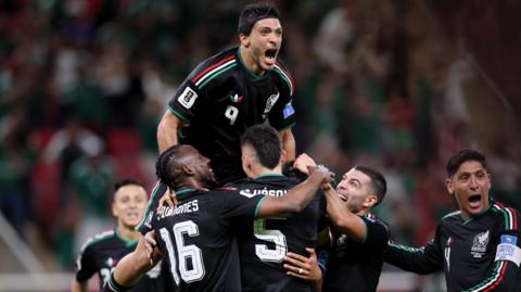 Raul Jimenez, wearing Mexico's black away shirt with red white and green Adidas stripes along the shoulders, celebrates their goal against South Korea at the World Cup with his team-mates by climbing on top of them. Edson Alvarez can be seen at the bottom right of the group of players with his mouth open celebrating.