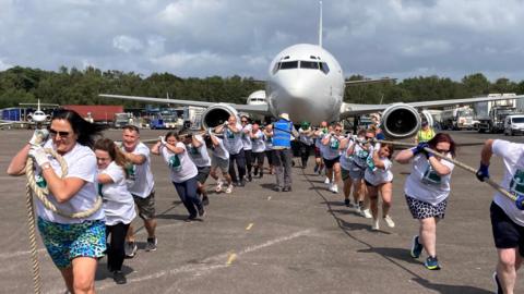 A group of people split in two are pulling a plane by two ropes on a slightly overcast day. A warden wearing a blue top and a hat can be seen in the middle of the two groups, in front of the airplane. Lorries and another plane can be seen in the distance.