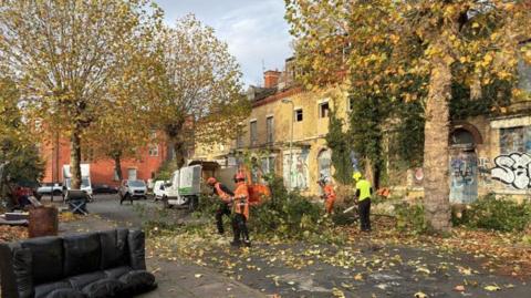 To the right of the image stands a row of derelict ochre-brick homes, with graffiti across the facade, boarded-up front doors and broken upper windows. Workmen in hi-vis jackets can be seen cutting back vegetation outside the terraced homes.  In the foreground, to the left, lies a discarded black sofa.