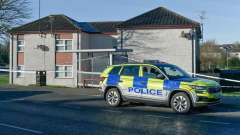 A white police cordon surrounds a block of flats in Corrainey Park in the Lisnahull Estate in Dungannon. It is a u-shaped, two-story building with grey render and panels of pink tiles surrounding its white windows. A blue and yellow police vehicle is parked in front of the cordon.
