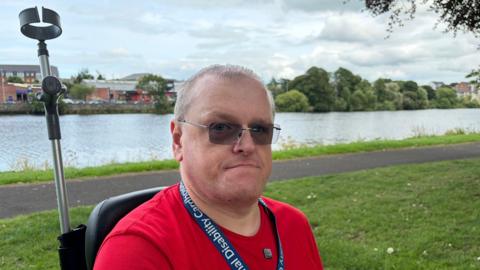 Stephen Magee has short grey hair and is wearing sunglasses, a red T-shirt and a blue lanyard. He is sitting in his mobility scooter in a park, with the River Bann visible in the background.