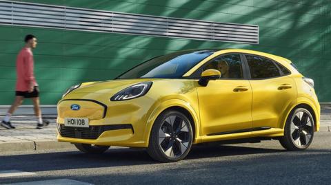 A bright yellow Ford Puma parked beside a street. A person in a red jacket, black shorts, and white sneakers walks on the pavement in front of a green building with horizontal white slats. The car faces right, and its license plate reads 'HOI108'.