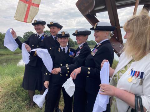 A group of six white women - five of which are wearing Royal Navy uniform, smile looking away from the camera and hold white handkerchiefs