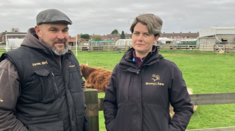 A man and a woman, both dressed in black jackets with yellow "Nunny's Farm" logos, stand in front of a brown Highland cow in a green field.