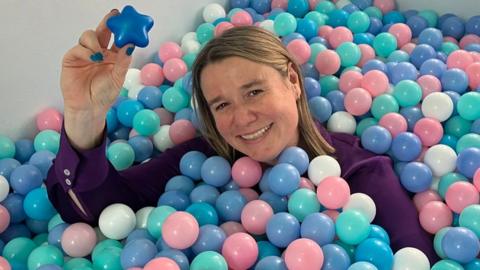Julie Williams smiles while partially submerged in a ball pit and holding up a blue plastic star