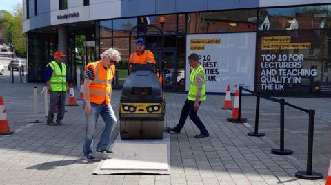 People in high visibility jackets stand by a road roller outside the University of Suffolk with wooden boards in front