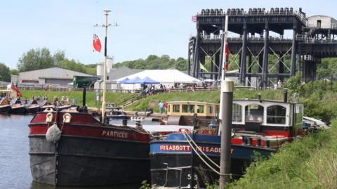 Boats and barges on a waterway with the massive boat lift structure behind and marquees next to it.
