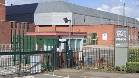 An industrial building behind a green fence and gate, with a gatehouse and a grey column outside with the Nestle logo on it and the words Nestle Confectionery