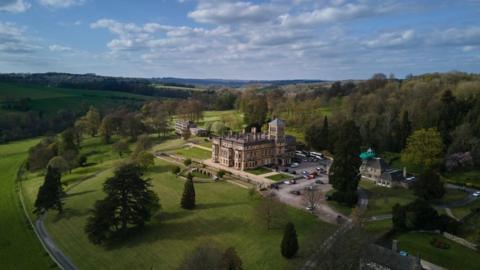 Drone photograph of a mansion, which serves as a private school, surrounded by parkland and woodland in the Cotswolds countryside. There is a car park to the side and manicured lawns surrounding the building.