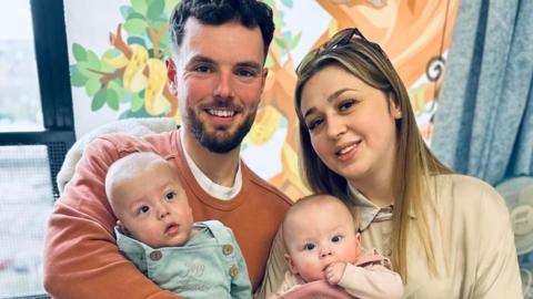 Cobie and Meghan standing together, each holding a baby. Cobie is standing on the left holding Joey who is dressed in a light blue outfit. Meghan is on the right holding Eva who is wearing a pink outfit and has her hand to her mouth. Behind them is a window and a colourful wall mural featuring illustrated animals and greenery.