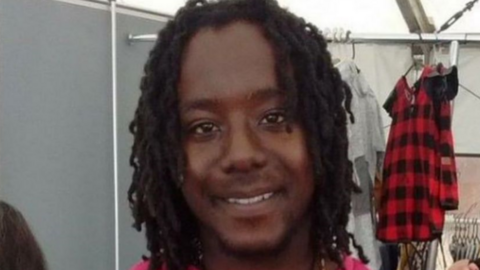 Curtis Green, a  young, black man with chin length hair who is smiling at the camera. Behind him are some clothes hanging off a clothes rail, including a red and black top.