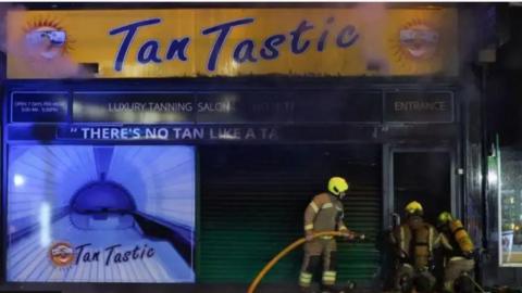 Three fire officers with hose outside exterior to shop window of TanTastic salon in Worthing