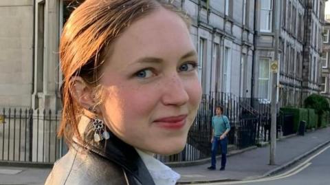 Rhiannon has brown, tied back hair and is wearing a black leather coat and white blouse. She is turned to the camera and is smiling. Behind her are a row of houses in Edinburgh where she was studying at university.