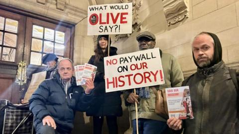 Five protestors from Save Our Museum outside Oxford Town Hall in November. They are holding signs which say "what would Olive say?!" and "No paywall on our history!"