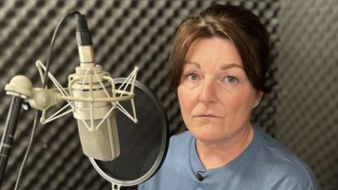 Gayanne is in a recording booth with black foam panels. She is sat beside her silver microphone which his hanging upside down with a black pop shield in front. She has brown hair, blue eyes and is wearing a blue top that says "The Bubbling Toad."