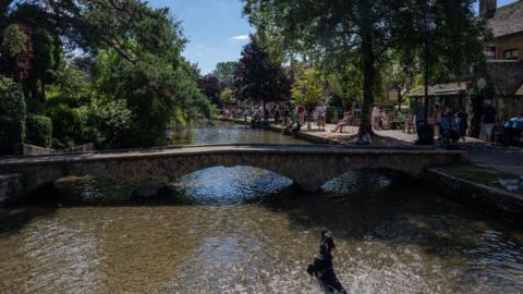 The River Windrush in Bourton-on-the-Water - a shallow river running beneath a low stone bridge. There are people standing along the river outside houses and there are trees growing on the banks. The sky is blue with very small white clouds.