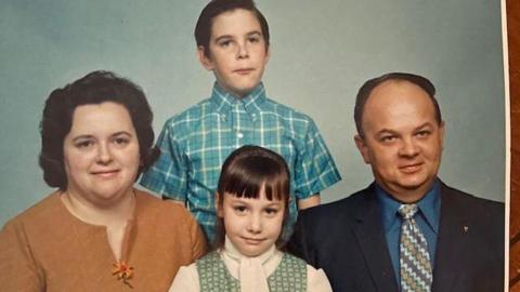 An old-looking colour photo of a man and a woman with their two adopted children, a son and a daughter. The mum has short dark hair and is wearing a brown cardigan, the dad balding and is wearing a blue and black suit, the son is wearing a blue checked shirt, and the daughter a green and white top.