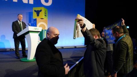 Side angle photo of protesters stood holding banners in front of Steve Reed, who can be seen in the background on the stage