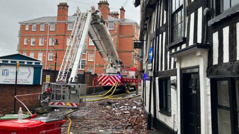 A red and white fire engine with a large silver aerial ladder and yellow hoses coming from the vehicle. There is a black and white building on the right and rubble all over the floor