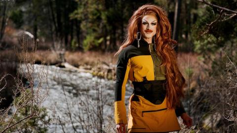 Performer Wyn Wylie, in drag as Pattie Gonia, wearing a yellow weatherproof dress with heavy face make up and long ginger hair. 