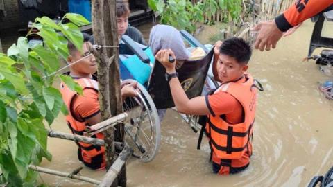 Philippine Coast Guard personnel rescue residents after flood waters rose from heavy rains brought by Tropical Storm Trami, in Bicol, Philippines, October 23, 2024. they are seen lifting a person in a wheelchair over the water