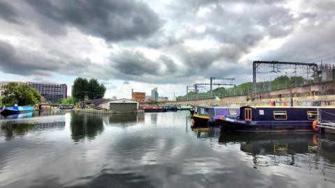 Grey shower clouds over a canal with two brightly coloured barges in the foreground and a railway bridge to the side