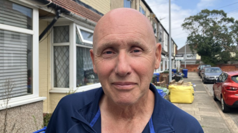 Bald man wearing a blue top looking at the camera. He is standing in front of terraced houses with cars parked along the street and wheelie bins behind him along the road.