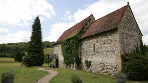 A small grey stone church. It has a brown roof and is in a field with a winding grey path leading to it