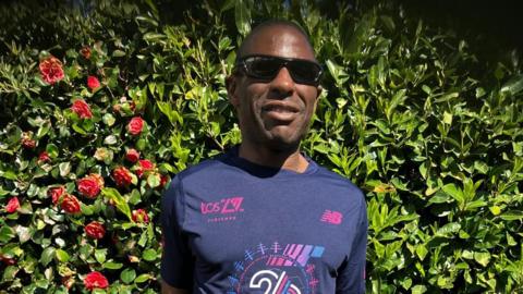 A man with sunglassed and a dark blue tshirt with purple writing is smilingat the camers as he stands against a bush with red roses on it