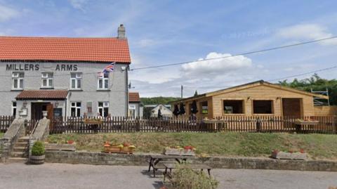 Outside of the pub with large wooden outdoor seating shed to the right. The pub is painted grey with an orange roof and matchung grey chimley. Around the two buildings is a brown painted fence. On the fence and below it are flower pots. There are four concrete steps leading up to the pub enterance.