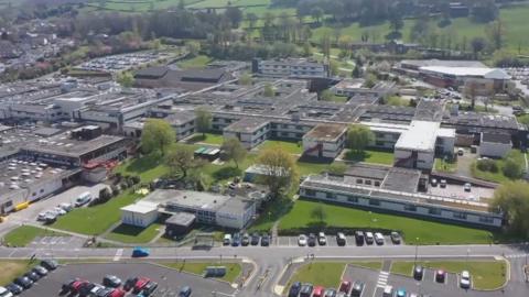 An aerial view of Airedale Hospital: car parks and green spaces can be seen in the foreground with an array of grey and white two-storey buildings behind. 