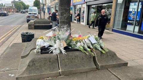 Flowers left around a tree in a street in Batley, West Yorkshire. There are several bunches of flowers, some with notes and ribbons on them.