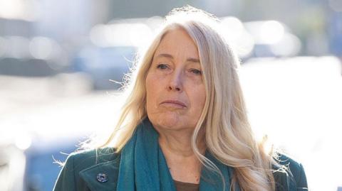 Sacha Dench is outside. It is a sunny day and sunlight is reflected on objects behind her. Dench has long blonde hair and is wearing a green coat and green scarf.