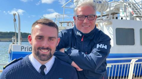 Head and shoulder shot of Stephen Jr and Stephen Snr - father and son. The younger man, to the left has a short dark hair, a close-cropped beard, and is wearing a dark blue v-neck jumper over a light blue shirt and dark tie. His father has grey hair, and is wearing spectacles and a navy blue jacket zipped up to the neck. Both are smiling and the Shields ferry can be seen just behind them.