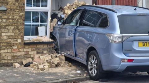 A picture of a light blue estate car that has crashed into the front of a house. The front of the car is buried into the front of the house next to a window. You can see rubble on the floor outside which is on the pavement.
