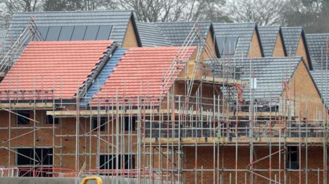 Houses under construction. Scaffolding is erected on the sides of brick houses. The buildings are incomplete and are missing windows.