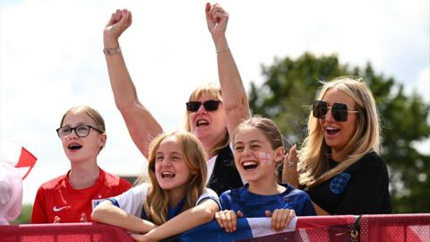 Five England fans smile at Southend Airport, waiting for the Lionesses to arrive home