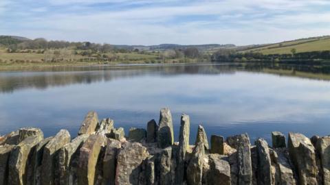 A rocky wall sits in front of a large still reservoir, with fields and trees in the distance on a sunny day.