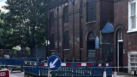A two-storey brick building featuring arched windows with metal grilles and white graffiti. In the foreground, blue road barriers and a directional traffic sign surround a cordoned-off pavement area. A side entrance displays a Star of David.