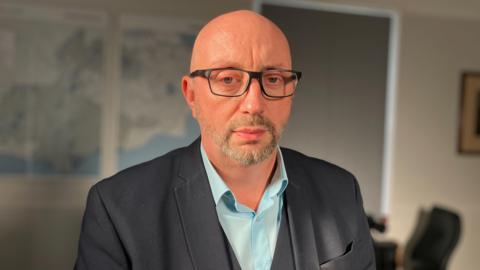 David Hinton - a man wearing a blue short and grey blazer. He has glasses, a stuble beard and no hair.