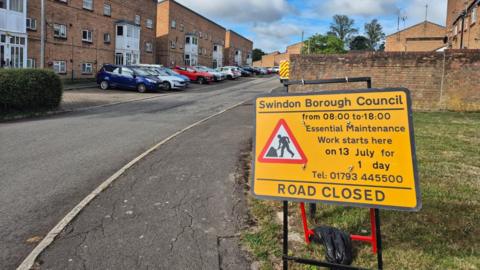 A bright yellow 'road closed' sign stands in the bottom right corner, in front of a residential road. Brick houses stand to the left, with a blue, white and red car in front.